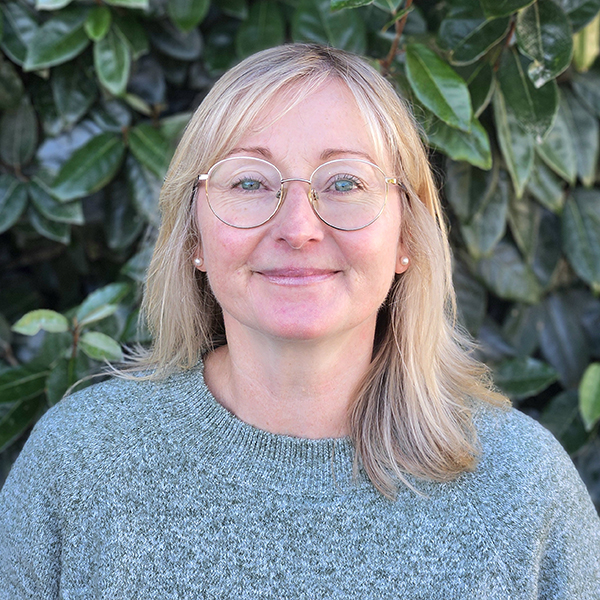 Jackie is dressed in a grey jumper, wearing glasses with foilage behind her.