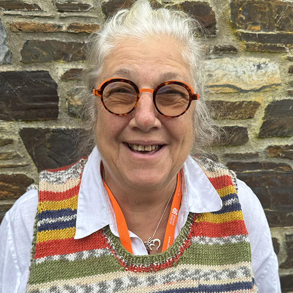 Lesley Stacy Marks, pictured wearing a colourful knitted tank top. It's striped with green, orange and yellow stripes. She is smiling and wearing round deep red glasses. Pictured against a brown stone wall