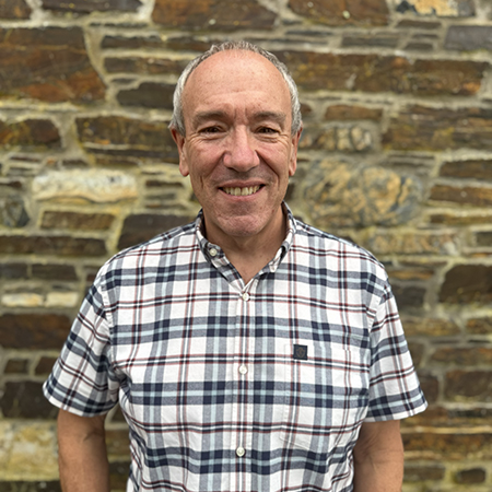 Suhail Shaikh is wearing a checked blue and white shirt. He is stood against a stone bricked wall.