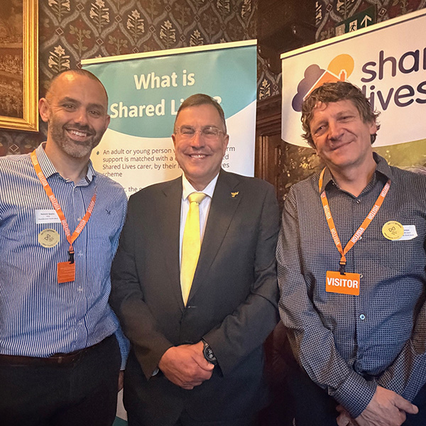 Shared Lives South West CEO Dominic Spayne is on the left, MP Martin Wrigley in the middle and Shared Lives Carer Ian Richardson on the right. They are photographed infront of two Shared Lives banners in parliament. Dom is wearing a blue shirt, Martin is in a suit with a lemon coloured tie and Ian is wearing a grey shirt.