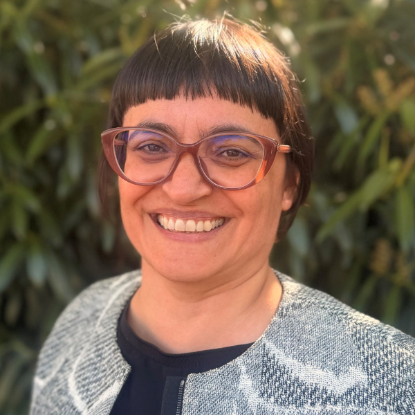 Sandra Fisher-Martins pictured smiling whearing a black and white jacket and mauve glasses. Pictured outside infront of greenery.